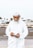 An elderly man with a white beard and traditional white attire is holding an open wallet. He is wearing a white cap and standing outdoors on a pavement with a road and vehicles blurred in the background.