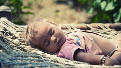 A baby is peacefully sleeping on a traditional woven cot outdoors. The child's eyes are closed and the expression is calm, with sunlight softly illuminating the face and the pink attire the baby is wearing.