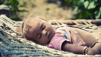A baby is peacefully sleeping on a traditional woven cot outdoors. The child's eyes are closed and the expression is calm, with sunlight softly illuminating the face and the pink attire the baby is wearing.