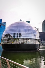 A futuristic spherical glass building floats on water, surrounded by modern skyscrapers. The reflection on the glass surface includes the surrounding architecture and features a prominent logo of a well-known technology company.