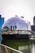 A futuristic spherical glass building floats on water, surrounded by modern skyscrapers. The reflection on the glass surface includes the surrounding architecture and features a prominent logo of a well-known technology company.
