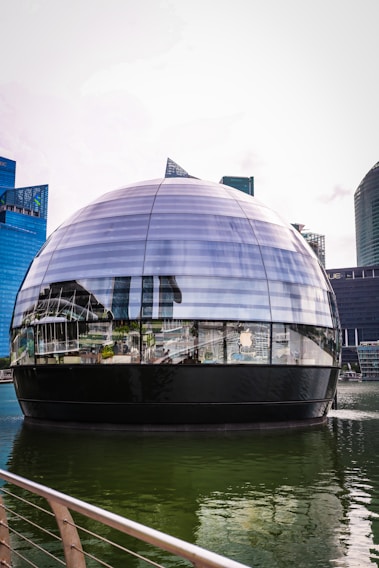 A futuristic spherical glass building floats on water, surrounded by modern skyscrapers. The reflection on the glass surface includes the surrounding architecture and features a prominent logo of a well-known technology company.