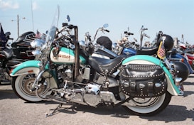 A detailed view of a shiny Harley-Davidson motorcycle with a classic design, featuring teal and white paint, leather saddlebags adorned with studs, and chrome detailing. It is equipped with a windshield and positioned among a row of other motorcycles. An American flag is attached to the rear of the bike.