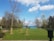 A serene landscape featuring a grassy area with newly planted trees staked for support. Mature trees border the scene, leading to a view of a large body of water under a blue sky with scattered clouds. A paved walkway runs along the grass, and a large urn planter is in the foreground.