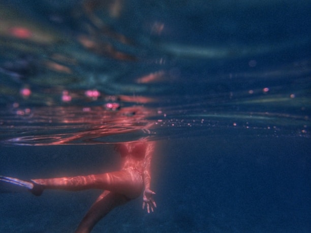 A person is swimming underwater, partially visible with their head just below the surface. The water is dark and illuminated with a mix of deep blue and red hues, creating a surreal and calm atmosphere. The light reflections and subtle bubbles add to the aquatic environment.