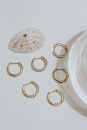 A collection of six small gold hoop earrings arranged on a light surface, accompanied by a textured white seashell. The lighting casts soft shadows, adding elegance to the minimalist composition.
