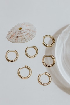 A collection of six small gold hoop earrings arranged on a light surface, accompanied by a textured white seashell. The lighting casts soft shadows, adding elegance to the minimalist composition.
