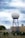 A large spherical radar dome mounted on a blue metal tower structure is situated in front of a beige building. Surrounding the structure is a fence with barbed wire, and various equipment is visible in the foreground. The sky above is cloudy with shades of blue and white.