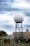 A large spherical radar dome mounted on a blue metal tower structure is situated in front of a beige building. Surrounding the structure is a fence with barbed wire, and various equipment is visible in the foreground. The sky above is cloudy with shades of blue and white.
