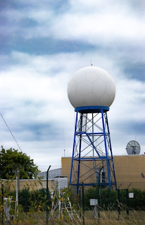 A large spherical radar dome mounted on a blue metal tower structure is situated in front of a beige building. Surrounding the structure is a fence with barbed wire, and various equipment is visible in the foreground. The sky above is cloudy with shades of blue and white.