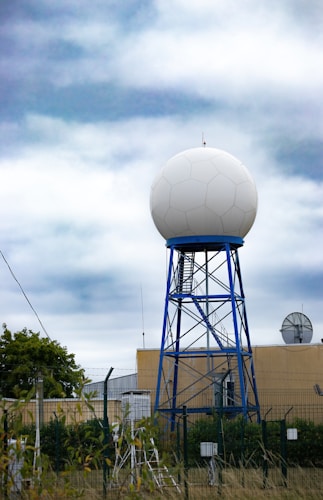 A large spherical radar dome mounted on a blue metal tower structure is situated in front of a beige building. Surrounding the structure is a fence with barbed wire, and various equipment is visible in the foreground. The sky above is cloudy with shades of blue and white.