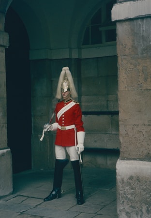 A soldier stands at attention in a formal ceremonial uniform, featuring a bright red coat with white accents and dark trousers. The soldier holds a sword and wears a highly polished helmet adorned with a white plume. The background consists of a stone wall and an arched doorway, suggesting a historical or official setting.