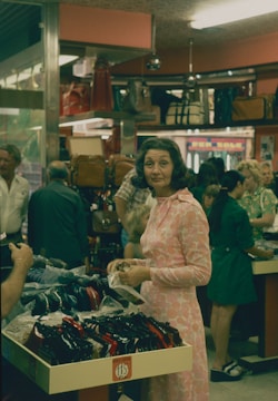 A busy retail environment with customers shopping for handbags. In the foreground, a woman in a pink dress holds a small item, while standing near a display of shoes or bags. Other shoppers and shelves filled with various types of bags are visible in the background, suggesting a bustling store.