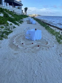 Two white-clothed tables with chairs are set up on a sandy beach. Each table is decorated with a center arrangement and surrounded by scattered flower petals arranged in circular patterns. The beach is bordered by greenery on one side and the ocean on the other. A modern building with a terrace is visible in the background.