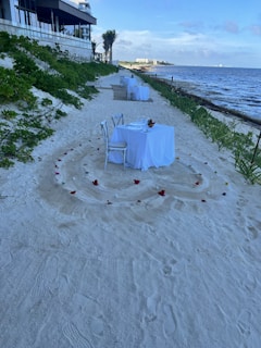 Two white-clothed tables with chairs are set up on a sandy beach. Each table is decorated with a center arrangement and surrounded by scattered flower petals arranged in circular patterns. The beach is bordered by greenery on one side and the ocean on the other. A modern building with a terrace is visible in the background.