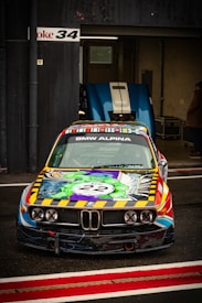 A colorful race car with a vibrant design featuring a comic book style. The car is parked in front of a garage, and another car with a blue hood is visible inside. The BMW Alpina logo is displayed prominently on the windshield.