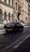 A street in a European city with classic architecture on both sides. The buildings have arched windows and ornate details. A black luxury car is driving on a cobblestone road lined with parked vehicles. The street is bathed in sunlight, casting shadows on the ground.