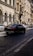 A street in a European city with classic architecture on both sides. The buildings have arched windows and ornate details. A black luxury car is driving on a cobblestone road lined with parked vehicles. The street is bathed in sunlight, casting shadows on the ground.