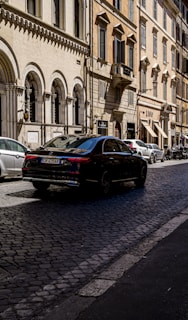 A street in a European city with classic architecture on both sides. The buildings have arched windows and ornate details. A black luxury car is driving on a cobblestone road lined with parked vehicles. The street is bathed in sunlight, casting shadows on the ground.