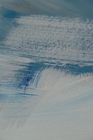 A close-up view of textured brush strokes in shades of blue and white, creating a smooth and layered appearance. The paint seems to be applied on a canvas, showing intricate details and variations in color and thickness.