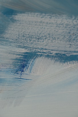 A close-up view of textured brush strokes in shades of blue and white, creating a smooth and layered appearance. The paint seems to be applied on a canvas, showing intricate details and variations in color and thickness.