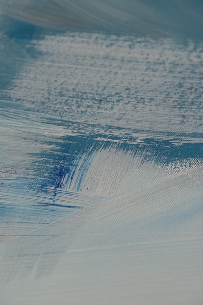 A close-up view of textured brush strokes in shades of blue and white, creating a smooth and layered appearance. The paint seems to be applied on a canvas, showing intricate details and variations in color and thickness.