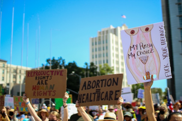 A group of people participates in a protest, holding signs advocating for reproductive rights and healthcare. The background features tall buildings and clear blue skies. Numerous signs have written messages such as 'Reproductive Rights are Human Rights,' 'Abortion = Healthcare,' and 'My Pussy My Choice.'