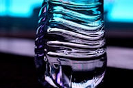 A close-up of a transparent, ribbed plastic bottle with light reflections creating an abstract pattern. The background is a gradient of blue, giving a cool and modern feel.