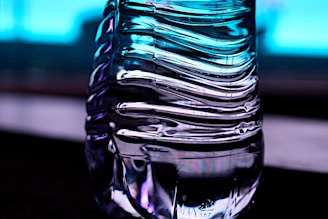 A close-up of a transparent, ribbed plastic bottle with light reflections creating an abstract pattern. The background is a gradient of blue, giving a cool and modern feel.