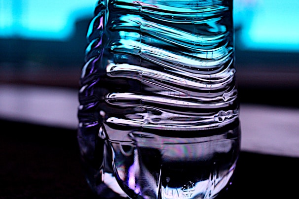 A close-up of a transparent, ribbed plastic bottle with light reflections creating an abstract pattern. The background is a gradient of blue, giving a cool and modern feel.