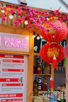 A vibrant and festive street food stall is decorated with red Chinese lanterns and artificial cherry blossoms. The stall features a menu written in what appears to be Russian and contains various food item names and prices. Behind the menu and decorations, there are shelves displaying jars and bottles, possibly containing sauces and condiments.