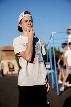 A young person wearing a white T-shirt and black shorts stands in an outdoor setting with a thoughtful expression, hand on their chin. The individual is also wearing a white cap backward. In the background, there are other people and some playground equipment.