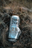 A can of Liquid Death with a design featuring the text 'Berry It Alive' is placed among dry, tangled branches and grass. The can has a shiny, metallic surface with intricate graphics.