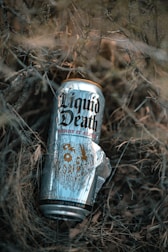 A can of Liquid Death with a design featuring the text 'Berry It Alive' is placed among dry, tangled branches and grass. The can has a shiny, metallic surface with intricate graphics.