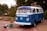 A vintage blue Volkswagen van is parked on a brick-paved path surrounded by greenery. Several retro items, including a crate with an old radio and steering wheel, are placed in front of the van. A luggage rack with additional items is visible on the roof.