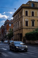 A dark-colored Mercedes-Benz car drives down a street lined with multi-story buildings. The buildings have an old European architectural style with a mix of yellow and red facades. There are other vehicles on the road, and the sky above is partly cloudy.