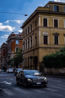 A dark-colored Mercedes-Benz car drives down a street lined with multi-story buildings. The buildings have an old European architectural style with a mix of yellow and red facades. There are other vehicles on the road, and the sky above is partly cloudy.