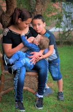 A woman is seated on a wicker chair outdoors, breastfeeding a baby. An older child stands close by, resting his head affectionately on her shoulder. The background features grass and trees, suggesting a natural setting.