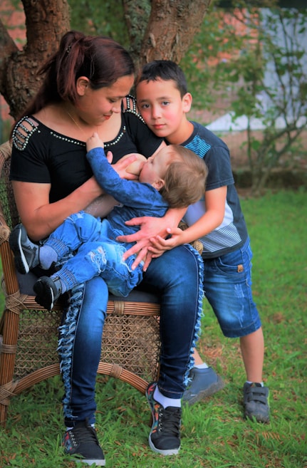 A woman is seated on a wicker chair outdoors, breastfeeding a baby. An older child stands close by, resting his head affectionately on her shoulder. The background features grass and trees, suggesting a natural setting.