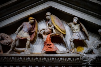 A stone relief sculpture depicting three muscular warriors engaged in battle. They are wearing ancient-style helmets and armor, with two holding shields and one appearing to be in distress. The figures are carved in high relief and have a dynamic, dramatic posture, suggesting a moment of intense conflict.