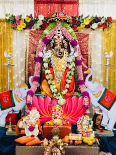 A richly decorated altar features a statue of a deity adorned with vibrant flower garlands and intricate jewelry. The background is adorned with sparkling red and gold fabric, flanked by floral arrangements. Two white elephants with traditional embellishments are positioned on either side. At the base, various offerings, including fruits, sweets, and small decorative items, are neatly arranged.