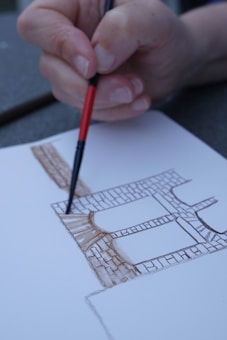 A hand holding a paintbrush is drawing architectural features on paper, using brown ink to outline the structure resembling a building with windows and stone textures.