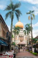 A vibrant urban scene featuring a prominent mosque with a large golden dome surrounded by palm trees. Shops and cafes line the path leading to the mosque, and people are casually walking and sitting in the area. The sky is bright blue with scattered clouds, adding to the lively atmosphere.