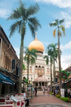 A vibrant urban scene featuring a prominent mosque with a large golden dome surrounded by palm trees. Shops and cafes line the path leading to the mosque, and people are casually walking and sitting in the area. The sky is bright blue with scattered clouds, adding to the lively atmosphere.