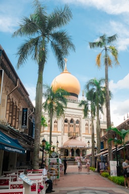 A vibrant urban scene featuring a prominent mosque with a large golden dome surrounded by palm trees. Shops and cafes line the path leading to the mosque, and people are casually walking and sitting in the area. The sky is bright blue with scattered clouds, adding to the lively atmosphere.