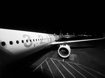 A monochrome photograph capturing an airplane on a tarmac during nighttime. The aircraft is prominently positioned with focus on the engine and wing. The fuselage displays large, partially visible numbers and letters, likely forming a promotional or identification message. The background shows dim airport lights and structures.
