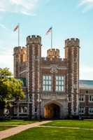 A historic university building with prominent stonework and architectural details, featuring towers and an archway entrance. Flags are flying atop the towers. The foreground includes a manicured lawn and pathway with a few people sitting.