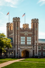 A historic university building with prominent stonework and architectural details, featuring towers and an archway entrance. Flags are flying atop the towers. The foreground includes a manicured lawn and pathway with a few people sitting.