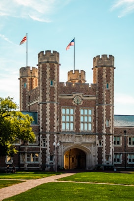 A historic university building with prominent stonework and architectural details, featuring towers and an archway entrance. Flags are flying atop the towers. The foreground includes a manicured lawn and pathway with a few people sitting.
