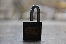 A padlock with a curved metal shackle is positioned against a blurred background. The padlock appears to be black with a metallic label on it.